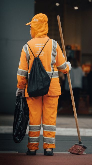 A street scene featuring a sanitation worker dressed in bright orange safety clothing, including a hooded jacket and reflective strips, engaged in surface cleaning. The worker is sweeping debris from 
