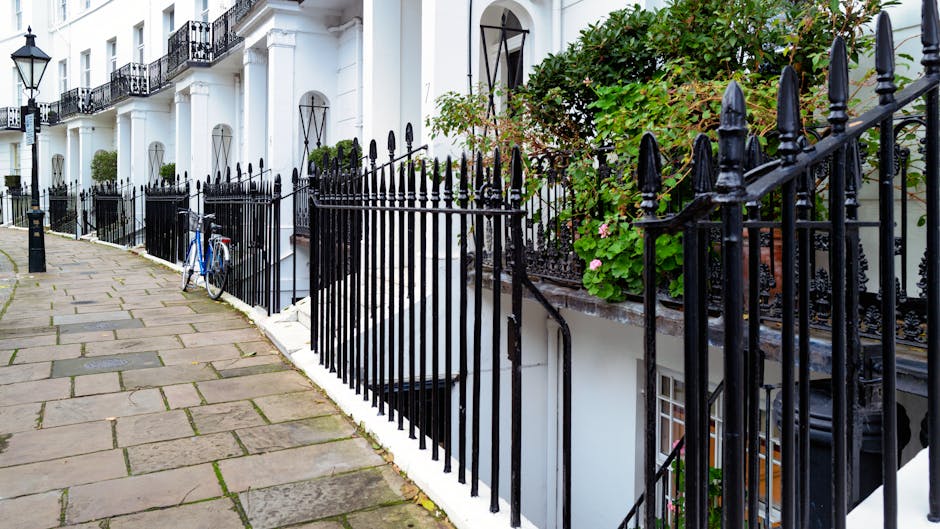 A narrow residential sidewalk with paving stones running alongside white terraced houses featuring black wrought iron railings. The railings encase small front gardens with green foliage and pink flowers, and some sections include downward stairs leading to basement flats. A blue bicycle is parked against one of the railings. The scene is well-lit with natural daylight, showcasing clean surfaces, freshly painted white walls, and well-maintained ironwork, reflecting a tidy, residential street in West Kensington. This image relates to professional domestic cleaning and maintenance services as discussed on West Kensington Cleaner’s webpage about access issues for basement flats cleaning.