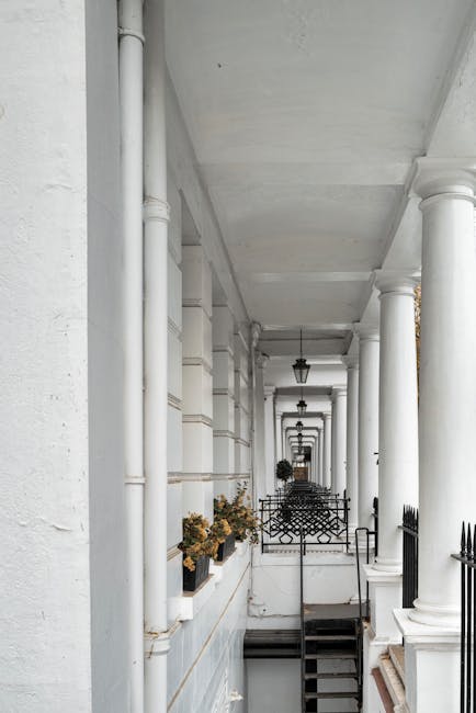 A covered residential corridor featuring white painted walls, large white columns supporting the ceiling, and black metal railings. The floor appears to be made of stone or concrete, with some decorative flower pots with plants placed along the wall. The area is illuminated by natural light and hanging lamps, giving a clean and well-maintained appearance consistent with professional cleaning standards by West Kensington Cleaner. The corridor shows no visible dust or dirt, and the surfaces are clean and smooth, indicating recent or ongoing surface cleaning and sanitisation activities relevant to basement flats access in West Kensington.
