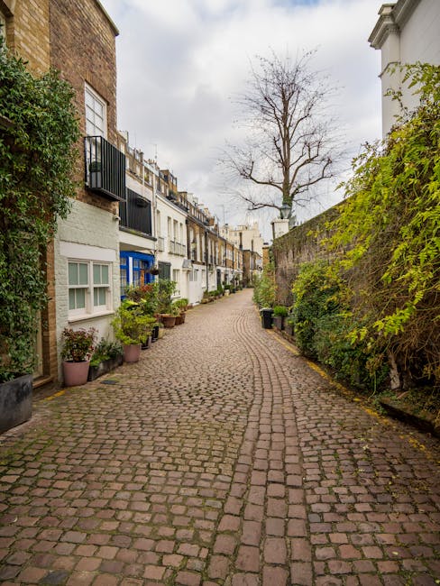 A narrow residential sidewalk with paving stones running alongside white terraced houses featuring black wrought iron railings. The railings encase small front gardens with green foliage and pink flowers, and some sections include downward stairs leading to basement flats. A blue bicycle is parked against one of the railings. The scene is well-lit with natural daylight, showcasing clean surfaces, freshly painted white walls, and well-maintained ironwork, reflecting a tidy, residential street in West Kensington. This image relates to professional domestic cleaning and maintenance services as discussed on West Kensington Cleaner’s webpage about access issues for basement flats cleaning.