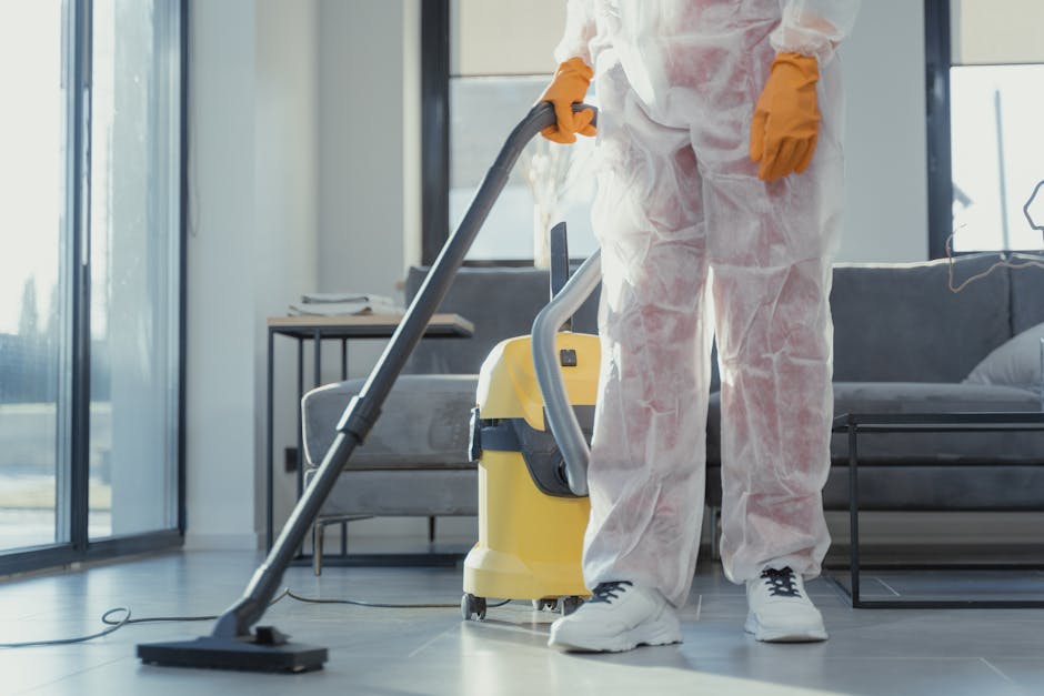 A professional cleaner wearing white protective coveralls and orange gloves is operating a yellow and gray vacuum cleaner during a deep cleaning session in a modern living room. The room features a light-colored wooden floor, a gray fabric sofa, and large floor-to-ceiling windows that allow natural light to illuminate the space. The surface of the floor appears dust-free and shiny, indicating thorough cleaning. Visible cleaning tools include a cloth and spray bottle on a nearby side table, with the vacuum cleaner’s nozzle directed towards the floor to remove dust and debris. The well-lit environment emphasizes the cleanliness and maintained condition of the room, which is part of a residential property receiving comprehensive cleaning by West Kensington Cleaner, specializing in deep cleaning and surface sanitisation for homes and businesses.