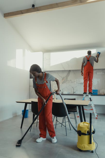 Two professional cleaners from West Kensington Cleaner are performing a thorough deep cleaning in a modern, open-plan kitchen and dining area. The room features a large marble splashback behind the countertop, which is being wiped down, and glossy ceramic tiles on the floor that appear spotless and shiny. One cleaner, a woman, is vacuuming the tiled floor with a yellow commercial vacuum cleaner, focused on removing dust from the surface. She is dressed in grey top and red overalls, and is working near a black dining table with black chairs. The other cleaner, a man, is standing on a step ladder, scrubbing or sanitising high-up areas on the wall or ceiling with a cloth, also wearing red overalls and a grey top, with overhead natural lighting flooding the space, highlighting its cleanliness and freshness. The scene emphasizes professional surface cleaning, deep cleaning, and sanitisation, showcasing West Kensington Cleaner's expertise in maintaining hygienic living environments in residential spaces.