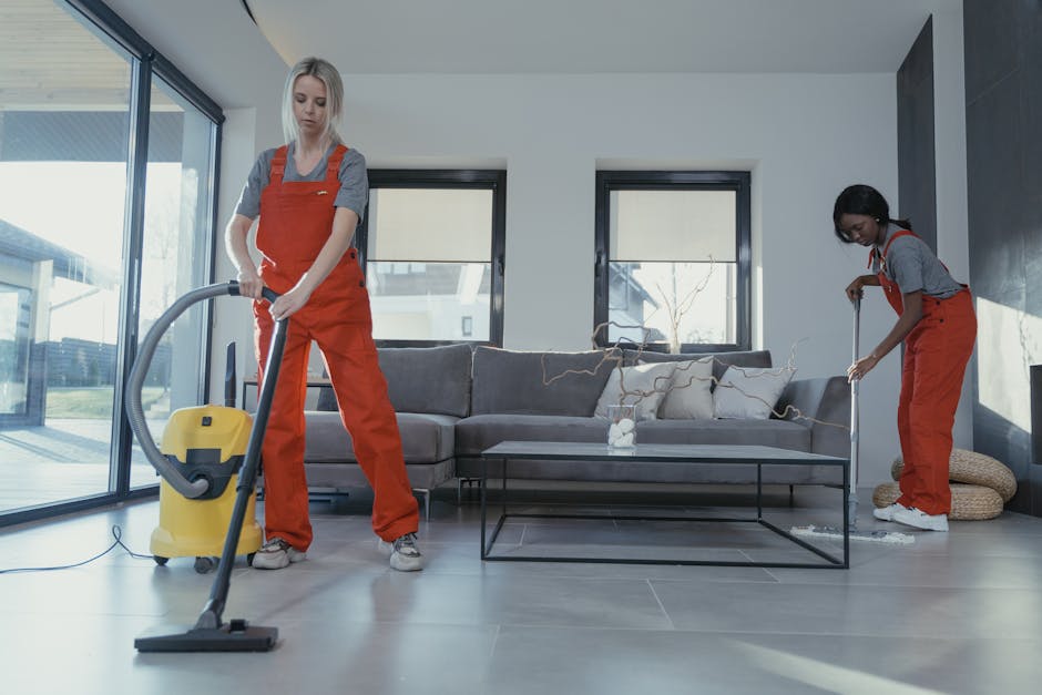 A professional cleaner wearing white protective coveralls and orange gloves is operating a yellow and gray vacuum cleaner during a deep cleaning session in a modern living room. The room features a light-colored wooden floor, a gray fabric sofa, and large floor-to-ceiling windows that allow natural light to illuminate the space. The surface of the floor appears dust-free and shiny, indicating thorough cleaning. Visible cleaning tools include a cloth and spray bottle on a nearby side table, with the vacuum cleaner’s nozzle directed towards the floor to remove dust and debris. The well-lit environment emphasizes the cleanliness and maintained condition of the room, which is part of a residential property receiving comprehensive cleaning by West Kensington Cleaner, specializing in deep cleaning and surface sanitisation for homes and businesses.