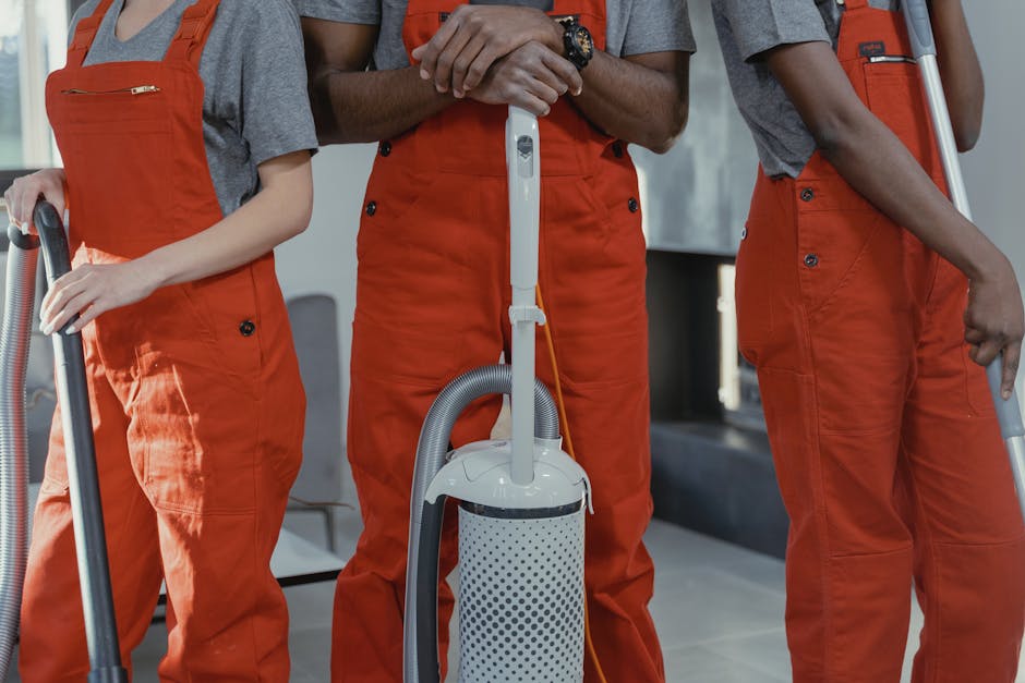Three professional cleaners from West Kensington Cleaner standing indoors, wearing grey t-shirts and bright orange overalls, each holding a vacuum cleaner with a grey hose and white body, ready to perform surface cleaning and deep cleaning tasks within a residential or commercial space. The background shows a modern room with tiled flooring and natural lighting, emphasizing cleanliness, hygiene, and maintenance activities associated with dometic cleaning services.