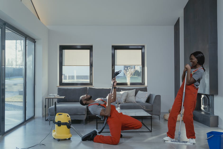 Three professional cleaners from West Kensington Cleaner standing indoors, wearing grey t-shirts and bright orange overalls, each holding a vacuum cleaner with a grey hose and white body, ready to perform surface cleaning and deep cleaning tasks within a residential or commercial space. The background shows a modern room with tiled flooring and natural lighting, emphasizing cleanliness, hygiene, and maintenance activities associated with dometic cleaning services.