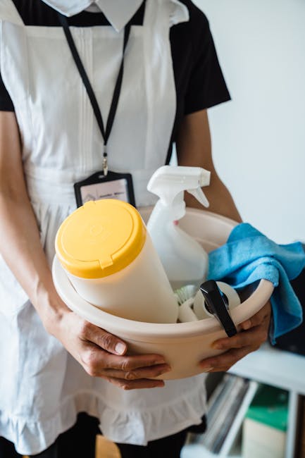A person dressed in a black and white uniform holding a white plastic container filled with cleaning supplies, including a spray bottle, yellow trigger spray, and a blue cleaning cloth. The background is plain and well-lit, emphasizing the tools used for surface and deep cleaning tasks. This image exemplifies professional domestic cleaning services provided by West Kensington Cleaner, focusing on hygiene and sanitisation for residential environments within Earls Court flats on Lillie Road.