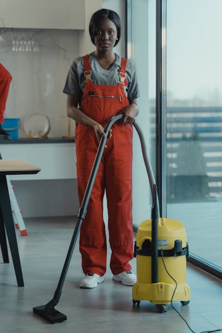 Two professional cleaners from West Kensington Cleaner, dressed in beige uniforms, standing back-to-back against a plain white wall. One cleaner, on the left, is holding a vacuum cleaner with a black handle and a red body, while the other, on the right, is holding a green-handled mop. Their uniforms are clean and pressed, indicating thorough preparation for domestic cleaning tasks. The scene showcases the tools used in surface cleaning and deep cleaning services, emphasizing hygiene and maintenance in residential or commercial environments. The lighting is bright and even, highlighting the pristine condition of the surroundings and the professional appearance of the cleaners, who are ready to perform sanitisation and detailed surface cleaning. This image exemplifies the high standards maintained by West Kensington Cleaner for flat cleaning services in Earls Court, Lillie Road, and West Kensington areas.
