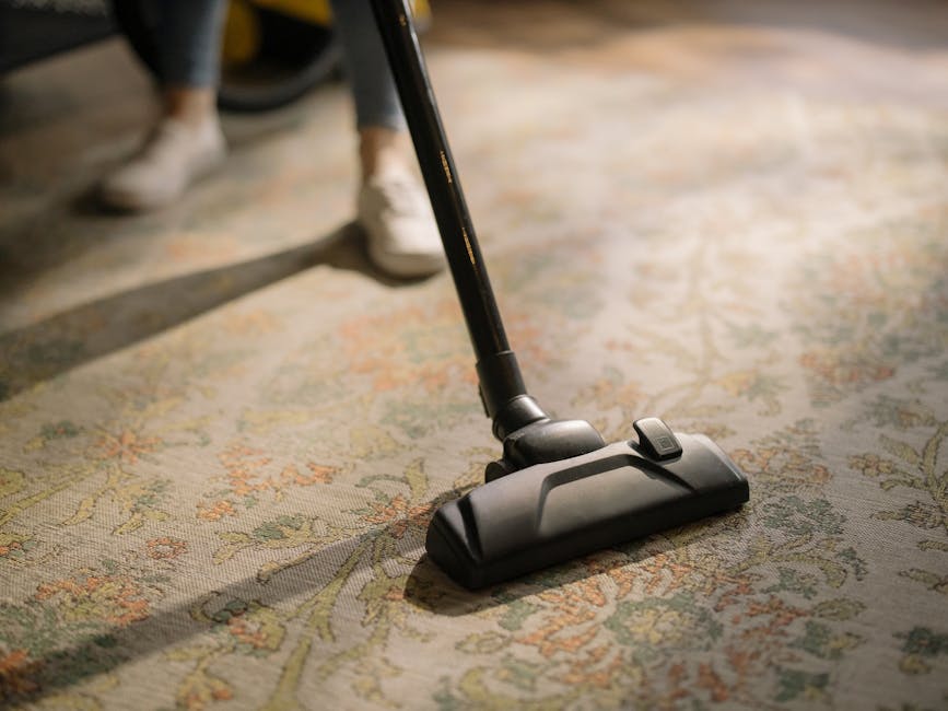 Close-up view of a vacuum cleaner head actively cleaning a traditional woven fiber carpet with intricate floral and geometric patterns in soft pastel colors. The vacuum is black and modern, with a fla