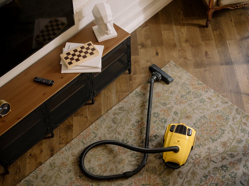 Close-up view of a vacuum cleaner head actively cleaning a traditional woven fiber carpet with intricate floral and geometric patterns in soft pastel colors. The vacuum is black and modern, with a fla