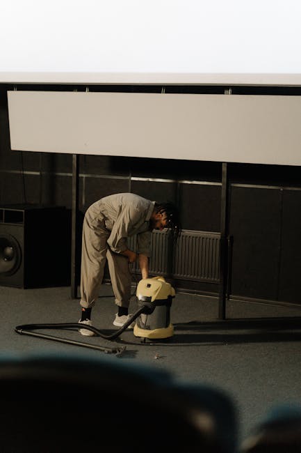 A person in a beige jumpsuit is engaged in deep cleaning a carpeted floor within an indoor space, likely an exhibition or conference venue, as indicated by the modern black wall panels and a white elevated structure in the background. They are using a yellow professional carpet cleaner with a hose attachment, focusing on a corner area near a radiator, which is mounted on the black wall. Visible around them are a large speaker or subwoofer, and the scene is well-lit with soft, even lighting highlighting the cleanliness of the area. This image exemplifies surface cleaning and maintenance appropriate for commercial or event spaces, aligning with services offered by West Kensington Cleaner for office and venue hygiene.