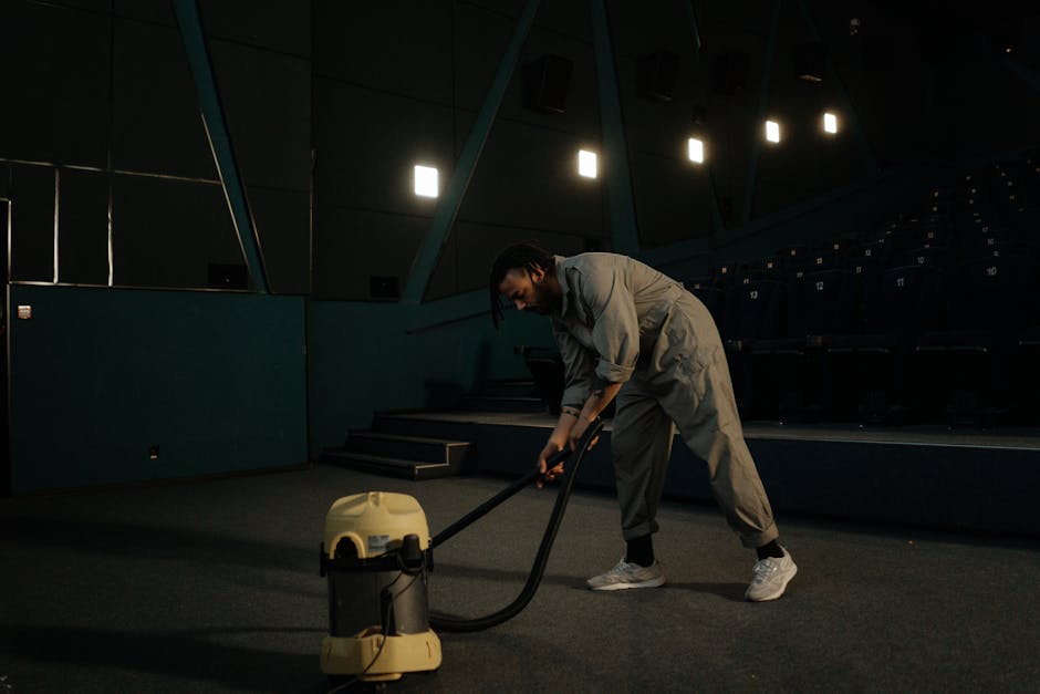 A professional cleaner at West Kensington Cleaner is performing deep surface cleaning in an indoor auditorium or conference room, vacuuming the carpeted floor with a yellow and black commercial vacuum cleaner. The room features dark walls with geometric paneling and stage steps at the back, illuminated by dim lighting from overhead fixtures. The clean, dust-free carpet appears well-maintained, and the overall atmosphere suggests thorough hygiene and maintenance typical of commercial cleaning services for event spaces or offices linked to Olympia London exhibitions.