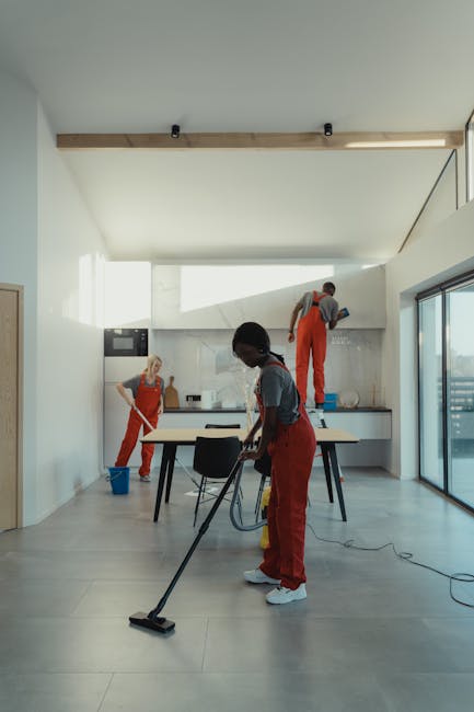 Image of three professional cleaners in a modern, bright kitchen with a minimalist design, engaged in surface cleaning and sanitisation. The room features a large floor-to-ceiling window that allows abundant natural light to illuminate the space. The floor is covered with large, light grey ceramic tiles, which appear freshly cleaned and shiny. One cleaner, dressed in red overalls, uses a vacuum or floor cleaning device on the tiled floor. Another cleaner, with dark hair and a grey top, is wiping or polishing a central white dining table, which is free of clutter. The third cleaner is positioned on a step ladder near the kitchen countertop, cleaning or inspecting the ceramic tiling behind the workspace. The kitchen cabinets are sleek, white, and handleless, contributing to the clean and modern aesthetic. This scene demonstrates domestic cleaning tasks performed by West Kensington Cleaner, ensuring hygiene and neatness in a contemporary residential setting, aligning with services highlighted in the page titled 'Same day cleaning West Kensington avoid hidden fees.'