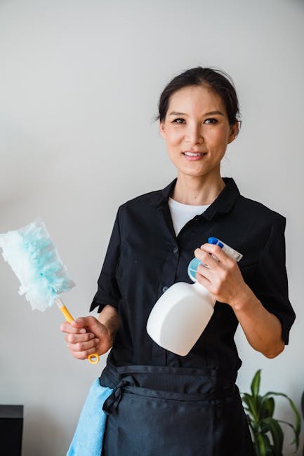 Two professional cleaners from West Kensington Cleaner, dressed in beige uniforms with the company logo, standing against a plain white wall in a well-lit room. One cleaner holds a red vacuum cleaner, while the other wields a green and white mop. The surfaces around them are clean and dust-free, suggesting thorough surface cleaning and domestic cleaning service. The environment appears bright with even lighting, emphasizing the cleanliness and hygiene of the space. The image highlights the expertise of West Kensington Cleaner in maintaining spotless, well-maintained surfaces, suitable for both residential and commercial cleaning needs.