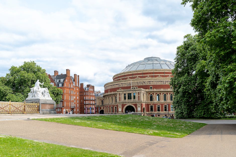 A large, historic building with a prominent domed roof constructed from red brick and stone accents, situated in a park-like setting with lush green trees and well-maintained grassy areas. The scene includes pathways made of gravel and paved surfaces, with a sculpture of reclining figures on the left side. The area appears clean and well-kept, with natural daylight illuminating the scene. The image showcases a public space likely used for cultural or civic purposes, captured during a partly cloudy day, highlighting the importance of surface and outdoor space maintenance, consistent with the cleaning standards discussed by West Kensington Cleaner on their website.