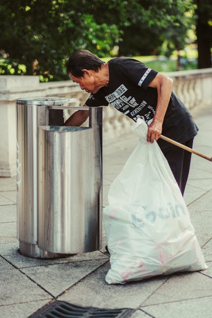 A woman with short dark hair, dressed in a black T-shirt, is seen bending over to dispose of trash into a cylindrical stainless steel litter bin with a horizontal opening, positioned on a paved outdoor area. She holds a white garbage bag in one hand and uses a stick or litter picker in the other to assist with waste collection. Surrounding her are lush green trees and a stone railing, with natural daylight illuminating the scene, emphasizing cleanliness and waste management practices consistent with urban maintenance and sanitation efforts. This image exemplifies routine outdoor cleaning and rubbish disposal, aligning with West Kensington Cleaner’s focus on effective surface cleaning and hygiene in public spaces.